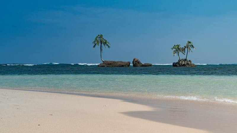 Plage de sable clair face à des rochers surmontés de cocotiers aux Cayos Zapatilla, dans le parc marin de Bastimentos au Panama, pays où se tourne Koh-Lanta All Stars. © Dronepicr, Wikimedia Commons (CC BY 3.0)