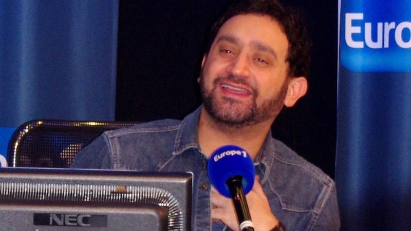 Cyril Hanouna souriant, la main sur la poitrine, devant un micro lors d'une émission de radio sur Europe 1 en 2014. © Julien Morvan / Wikimedia Commons (CC BY-SA 3.0)