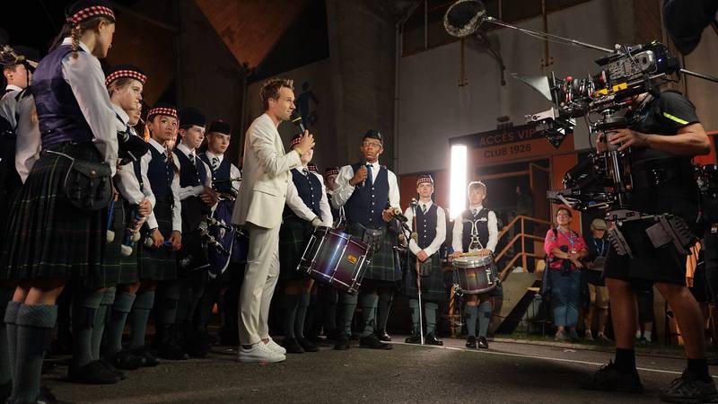 Cyril Féraud en costume blanc, micro à la main, devant un orchestre de cornemuses en kilt lors d'un tournage au Festival interceltique de Lorient en 2024